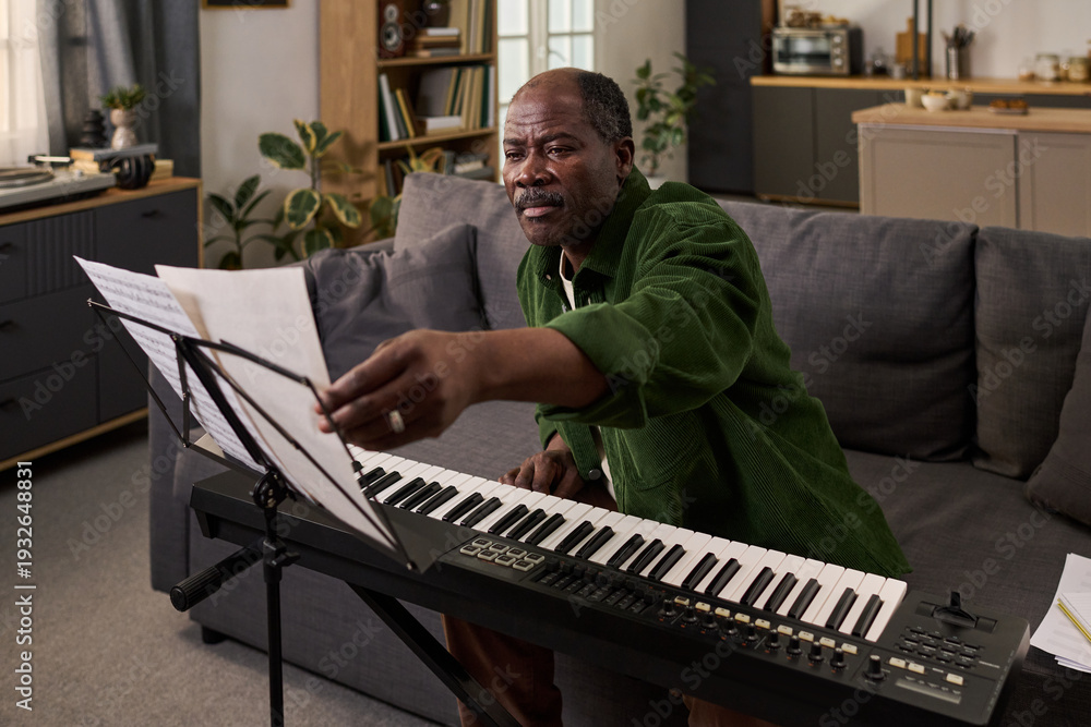 Fototapeta premium Middle aged Black man sitting on sofa playing digital piano, reaching for sheet music on music stand, practicing music in modern living room with focused expression