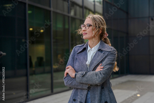 Quadro em tela Young woman stands outside a modern building with arms crossed while looking awa