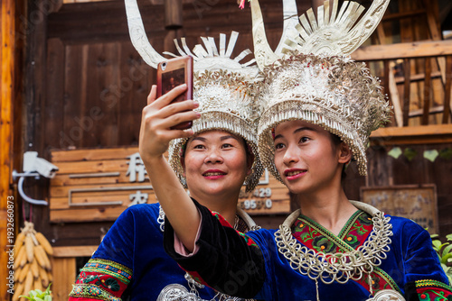 China, Guizhou, two Miao women wearing traditional dresses and headdresses taking a selfie with smartphone