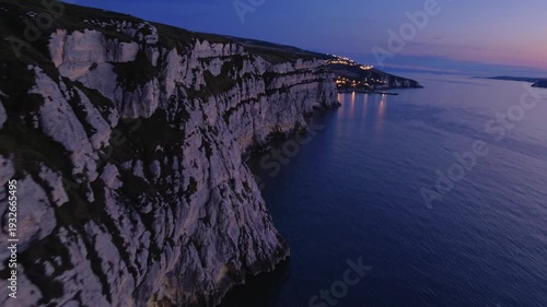 Aerial view of rugged coastal cliffs illuminated by twilight, showcasing the serene ocean and distant lights of a coastal town along the shoreline
