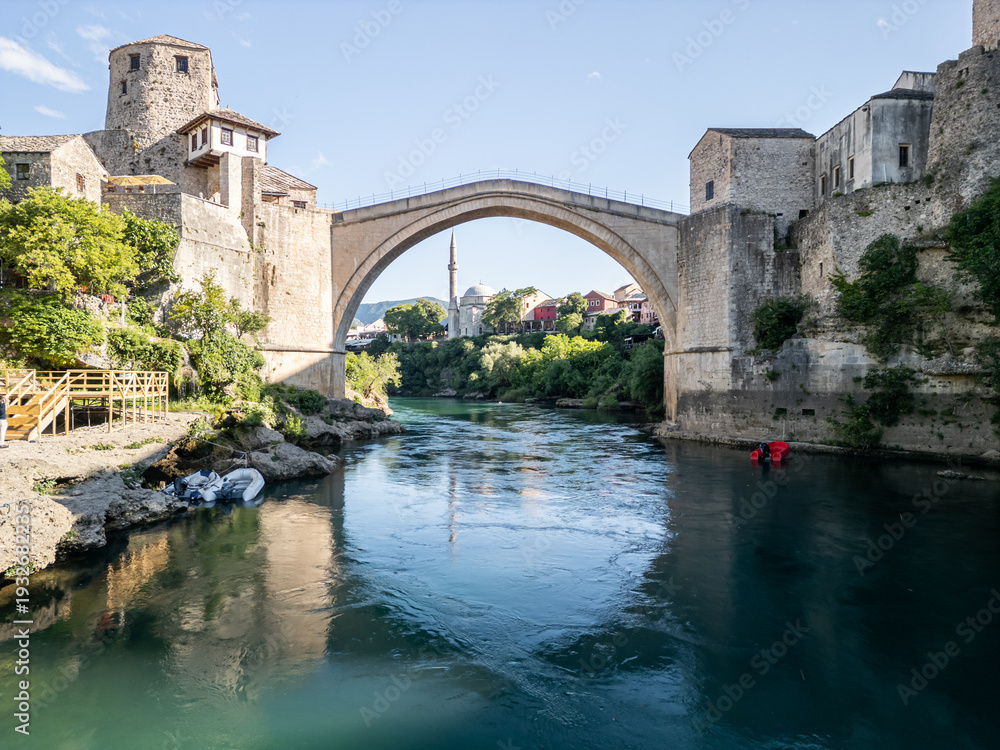 Obraz premium Mostar, Bosnia and Herzegovina 05 28 2025: Frontal drone photo of Old Bridge Mostar in Old Town with Halebija Tower, Sultan Selimov Mesdžid, Koski Mehmed Pasha Mosque, Neretva River, boats, and Beach