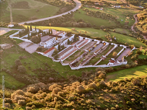Aerial view of a cemetery with white buildings and rows of graves surrounded by lush green hills, Manilva, Spain.