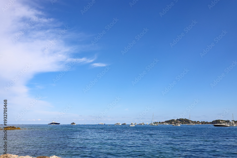 Obraz premium Calm blue waters of the Mediterranean sea with distant yachts near Beaulieu-sur-Mer, Peaceful seascape at the French Riviera with rocky coast and wide open sky