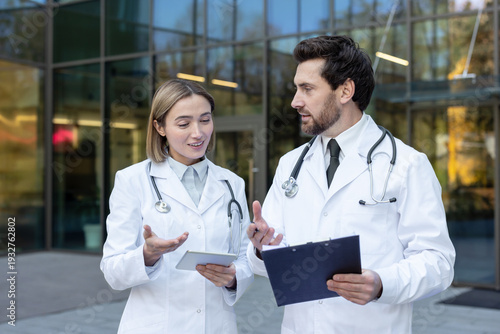 Two doctors confer outside a modern clinic, reviewing a tablet and clipboard as they discuss patient care, diagnosis and treatment plans in a professional teamwork setting