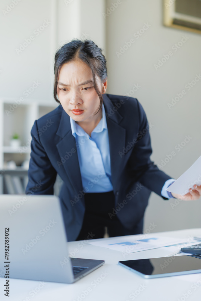 Naklejka premium Asian businesswoman concentrating with a confused expression, leaning over a laptop while holding documents, experiencing stress and a problem at work in the office