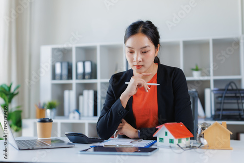 Businesswoman evaluating real estate investment and mortgage options at a modern desk, laptop open with house models, calculator and documents for property planning and financing decisions
