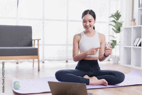 Young woman sitting in lotus position on a yoga mat with a laptop, pointing at a glass of water while participating in an online fitness class at home Free Download Young woman sitting in lotus position on a yoga mat with a laptop, pointing at a glass of water while participating in an online fitness class at home - GFXLoad