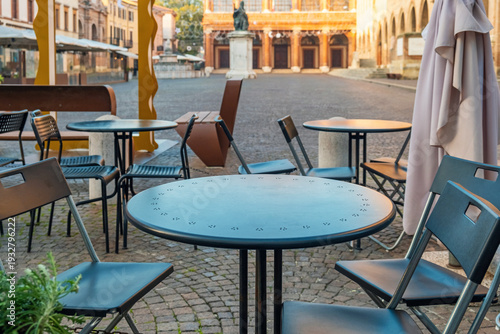 Outdoor cafe tables and chairs sit on cobblestones in Piazza Cavour, Rimini, Italy, with statue and historic civic buildings in the background. Italian street restaurant with nobody at sunrise