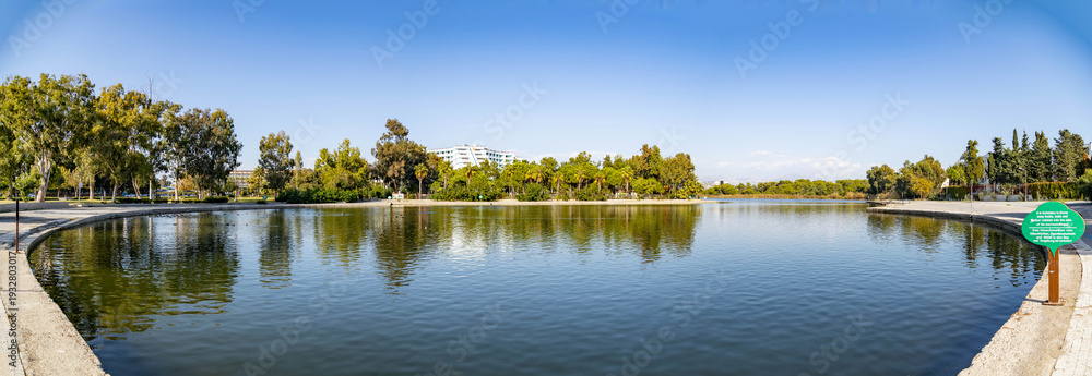 Fototapeta premium Panoramic view of a calm lake with tree reflections in Sorgun Titreyengol. Scenic landscape with green foliage, blue sky, and hotel buildings in Antalya province, Turkey. Sunny autumn day.