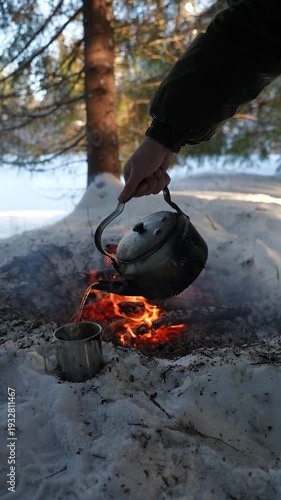 Steaming hot tea is poured from a camp kettle into a metal mug resting on fresh snow. Winter camping in the backcountry. A warming drink during a cold-weather hike. Bushcraft, outdoor cooking, and