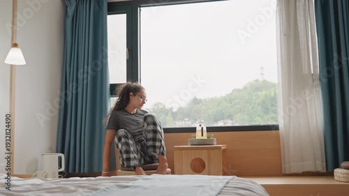 Guest in hotel room morning young woman moves to window sits on bench sets cup on wooden table gazes at green skyline beyond soft natural light through blue curtains bedside lamp neatly made bed calm