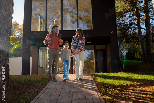 Smiling family of four, including parents with a young boy on father's shoulders and a girl holding mother's hand, walking on a paved path in front of a contemporary home during autumn