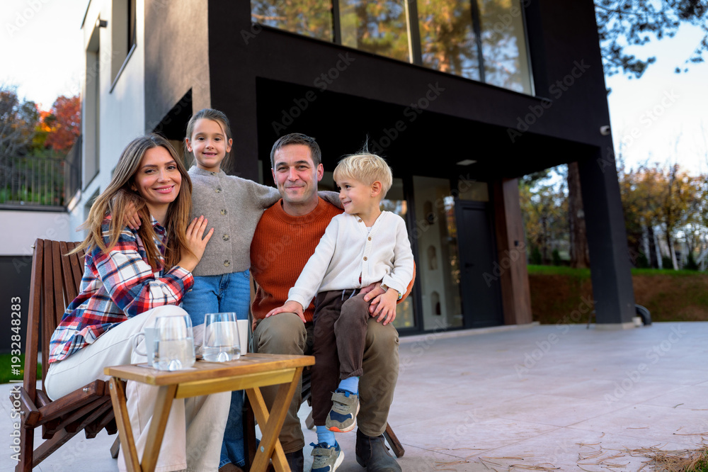 Fototapeta premium Parents with their young children smiling and posing for the camera, sitting on a private terrace of their modern house, representing family lifestyle and togetherness