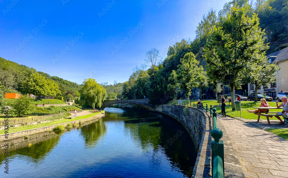 Fototapeta premium Esch-sur-Sure, Wiltz, Grand-Duche de Luxembourg, September 07, 2025, sunny river scene with stone bridge and riverside seating, cheerful blue sky and people-friendly promenade where local artist