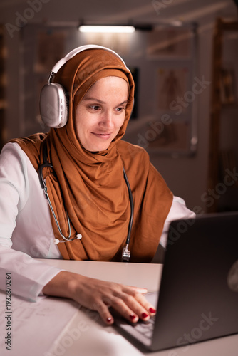 Female Muslim doctor in hijab and white coat using headphones and laptop computer during telemedicine session on night shift at hospital. Medicine, healthcare and people