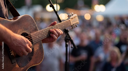 Close-up hands strum an acoustic guitar on stage with a microphone and colorful crowd bokeh in the background, creating a warm energetic live music atmosphere.