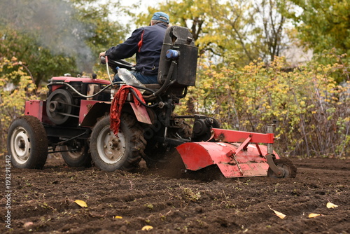 Billede på lærred Man on red tractor plowing the field, side-back view, smoke from chimney, horizontal shot
