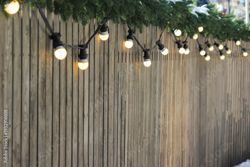 Cozy festive background with warm glowing garland lights on a wooden fence decorated with fir branches.