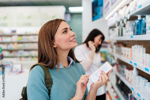 A young woman examines the ingredients on a medicine box while shopping in a modern pharmacy with a pharmacist working in the background.