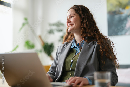 A young woman with curly brown hair smiles while working on her laptop in a bright, modern office setting.