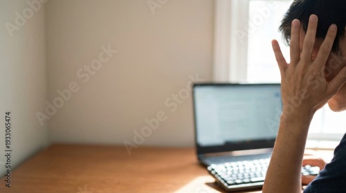An individual with hand covering face appearing stressed or overwhelmed working on a laptop at a sunlit desk