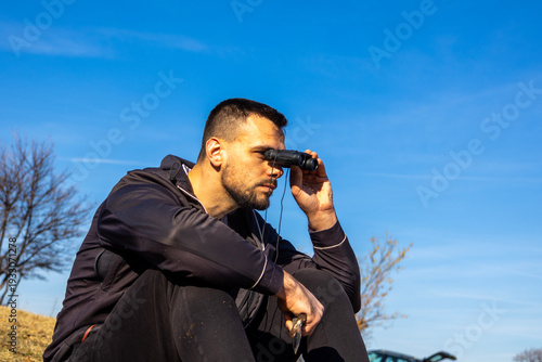Obraz na plátně Young man seated on a dry grassy hill using binoculars to observe the distance