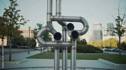 A sculpture of industrial metal pipes stands in a park with a cityscape in the background at daytime.