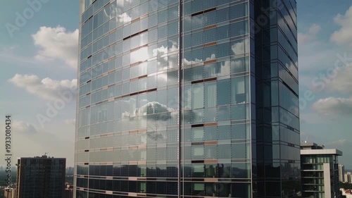 A sleek glass skyscraper reflects clouds and cityscape on a sunny day with modern architecture nearby