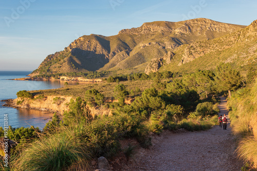 Hikers walking on a coastal trail at Playa Es Calo in Mallorca during golden hour with mountain views