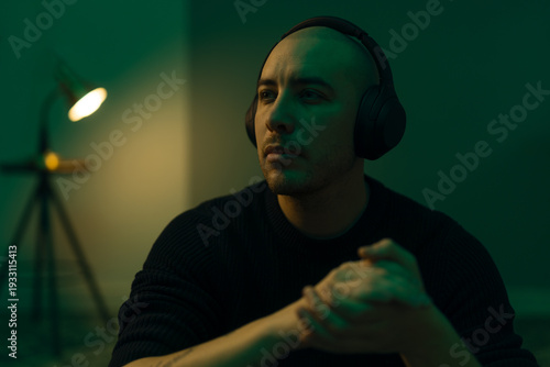 Pensive young man with headphones sitting in a moody, green-lit room. Cinematic portrait of a thoughtful person listening to music or a podcast in a dark, atmospheric interior with warm lamp light.