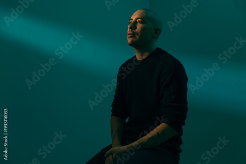 Thoughtful man with tattoos sitting in a dark, moody studio. Dramatic teal lighting with a sharp diagonal light beam creates a cinematic, contemplative atmosphere against a deep green background.