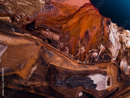 Aerial view of the striated earth unfurls in bands of deep umber and sun-baked ochre, carved by time and industry, Aluminium Refinery, Western Australia, Australia.