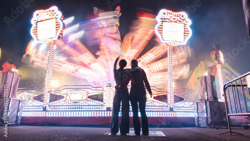 Fotografie Two friends embrace, watching the bright, spinning carnival ride at fairground