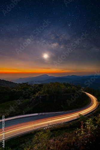 Long exposure of car light trails on a winding road at Doi Inthanon, Thailand. Stunning night landscape with the Milky Way galaxy, moon, and orange twilight glow over mountain ranges.