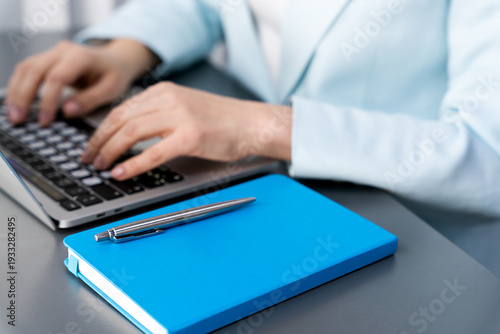 Modern business workspace concept highlighting productivity, organization and digital workflow. Blue notebook and pen on desk and in the background are the hands of a woman working on a laptop.