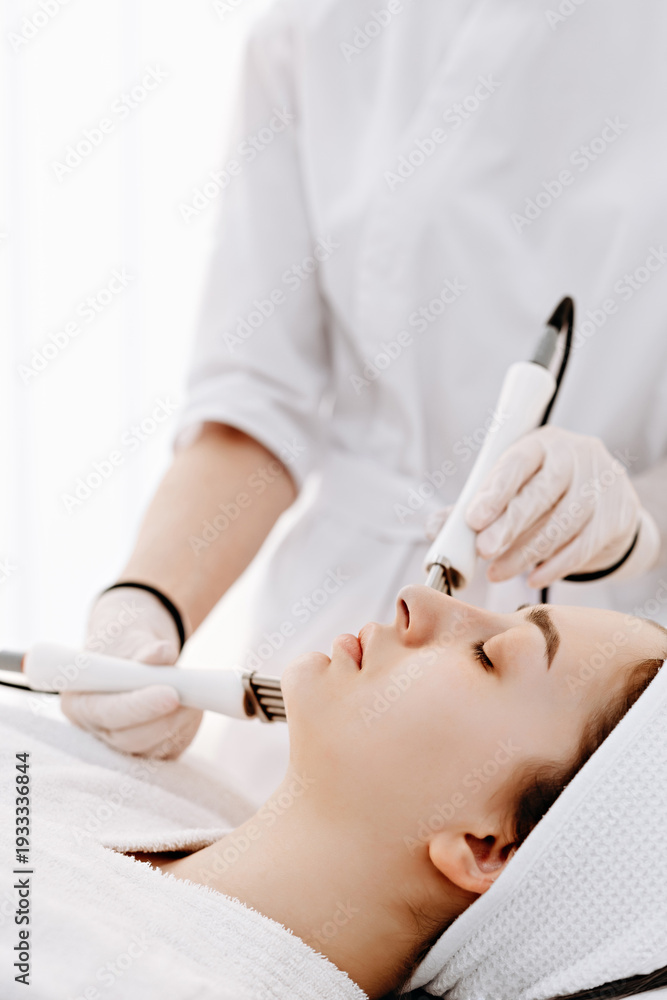 Naklejka premium Woman receiving professional facial microcurrent treatment at spa by aesthetician in white uniform