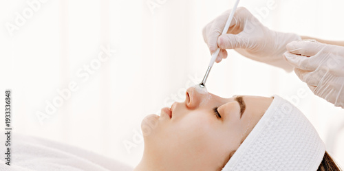 Woman receiving professional facial treatment at spa as esthetician applies skincare mask to her nose