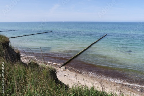 In Ahrenshoop auf der Halbinsel Fischland wachsen Gräser an der Steilküste. Unten sind Buhnen in der Ostsee zum Küstenschutz. Das Meer ist ruhig an diesem Sommertag.