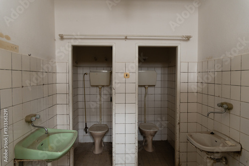 Interior of an abandoned institutional bathroom with two toilet cubicles, old pedestal toilets with high cisterns, and a green ceramic washbasin on the left