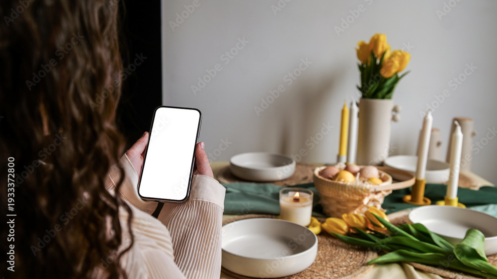 Fototapeta premium Woman holding a smartphone with a blank screen over a festive Easter table setting