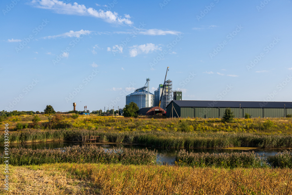 Naklejka premium Golden fields stretch around a serene pond, with a grain storage facility and cranes rising against a clear blue sky, capturing rural life