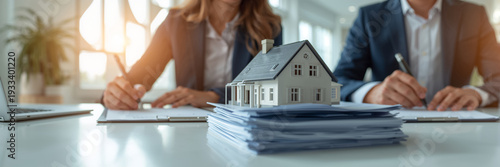 There's a model of a house on the table, and a realtor and a buyer are signing a sales contract nearby.