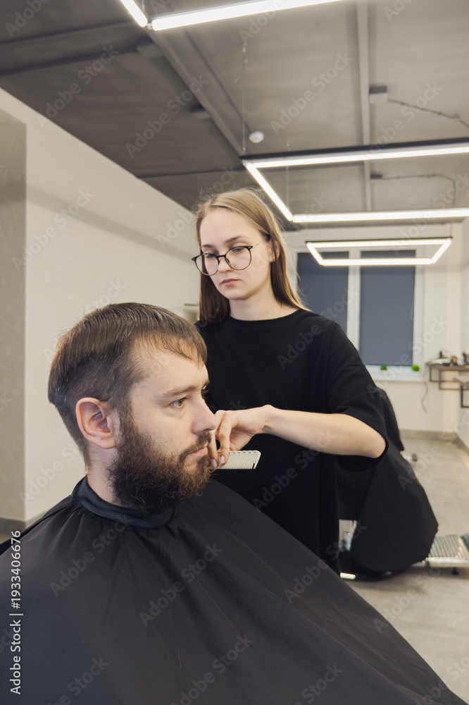 Fototapeta premium Professional woman barber wielding electric hair clipper making Fashionable haircut. Bearded Man client visit gentleman grooming beauty salon for hairstyle care. Modern Haircut style in barber shop