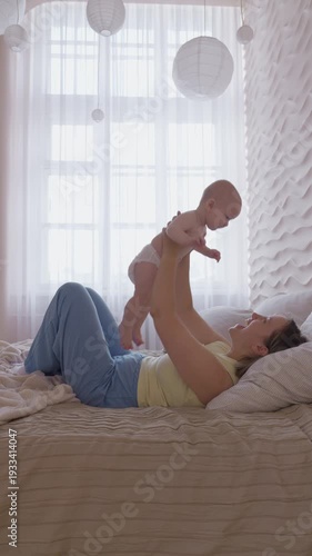 mother lifting baby above chest on bed. morning exercise and play mom with infant with bright window light. cheerful movement and eye contact. happy domestic maternal routine. cozy bonding