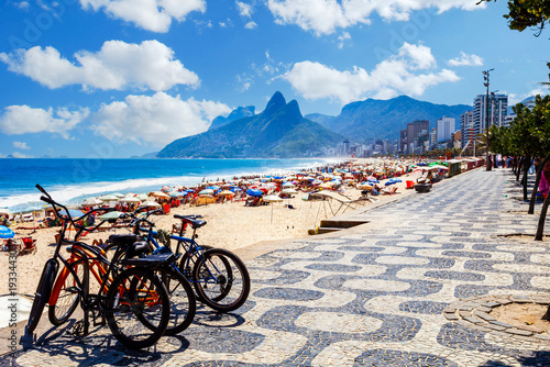 Fotografie Sunny panoramic view of Ipanema Beach in Rio de Janeiro, Brazil, featuring the iconic wavy mosaic promenade, parked bicycles, and the Morro Dois Irmãos mountains under a blue sky