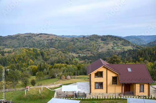 Modern wooden house on a hillside overlooks forested mountains and rolling green valleys, conveying peaceful rural living, nature, and open-air scenery beneath a soft overcast sky. Carpathians