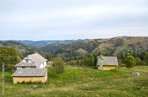 Wooden farmhouses and outbuildings rest on a grassy hillside overlooking a forested valley and rolling hills, evoking peaceful rural life, pastoral scenery and an idyllic mountain countryside retreat