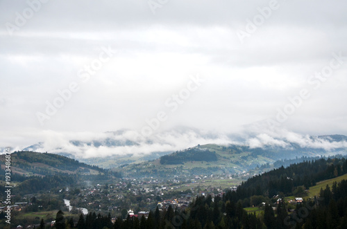Misty mountain Verkhovyna village nestled in a forested valley, with low clouds drifting over rolling hills and scattered houses, creating a peaceful, moody landscape scene. Carpathian Mountains