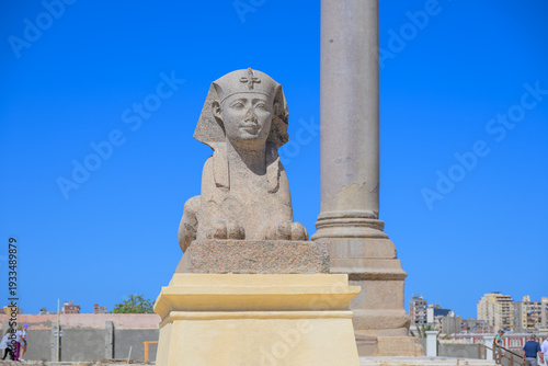Фотография Front view of the pink granite Sphinx with Pompey's Pillar behind at the ancient Serapeum Complex in Alexandria, Egypt