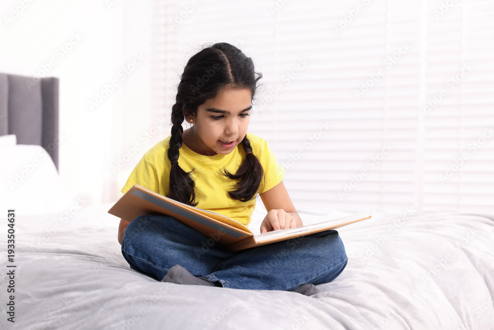 Naklejka premium Cute little girl reading book on bed at home